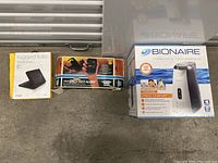 Lot showing ZAGG Rugged Folio box, Ansmann battery pack, Bionaire humidifier box, and another boxed electronic item, all on a metal surface