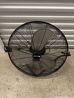 Black metal oscillating floor fan on concrete floor with protective grille and three blades visible. Power cord laid out.