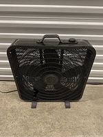 Black plastic box fan on concrete floor in front of a metal shutter door, displayed upright on two feet with power cord visible.