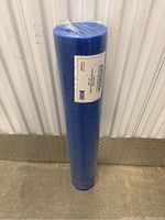 Image of a single blue foam roller standing upright, wrapped in plastic, placed against a white wall with a metal strip along the bottom.