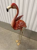 Side view of red metal flamingo statue showing detailed feathers, beak, and legs, standing against a metal wall base.