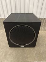 Front view of black cube-shaped Polk Audio powered subwoofer with round speaker grille on front.