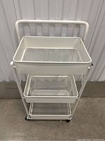 Front view of the white metal rolling cart showing three mesh baskets and caster wheels