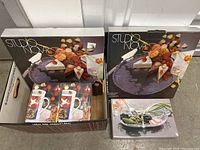 Top view of box containing two Studio Nova serving plates in original packaging and two boxed Christmas mugs.