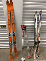 Full view of two pairs of skis and two pairs of ski poles leaning against a wall.