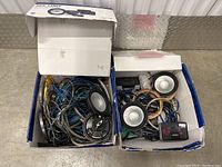 Two open cardboard boxes side by side containing assorted speaker parts and cables, photographed from above showing tangled wires and speakers.