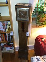 Full view of the tall wooden antique grandmother clock next to bookshelf and window. Shows overall height and design of the clock.