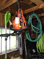 Front view of orange Echo chainsaw hanging from chain in shed alongside green electric tool and garden hoses