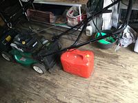 Wide shot showing green push mower with black grass catcher and red fuel can on wooden floor