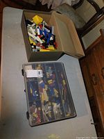Cardboard box filled with assorted LEGO pieces and plastic storage bin with compartments filled with assorted LEGO parts on a table.