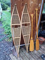 Photo showing full view of two vintage wooden snowshoes next to two wooden canoe paddles against a wooden wall