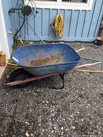 Front and side view of the YardWorks wheelbarrow showing blue metal bin, wooden frame, and black rubber tire on gravel ground near a blue building.