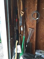 Yard tools including axe, saw, rake, steel bar, pole pruner, and wire coil against barn wall.