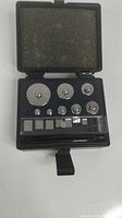 Closed black plastic case containing a set of metal calibration weights organized by size with foam lining inside lid.