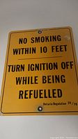 Front view of vintage yellow metal sign with black text stating no smoking and ignition off instructions during refueling.