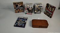 All items displayed together on a white surface: two Grey's Anatomy DVD sets, two Funko Pop! figures in boxes, Xbox game case, and brown vintage purse.