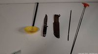 Photo showing five tools including a yellow plastic fishing skimmer ladle, hunting knife with sheath, sharpening rod, and a long metal rod with red handle.