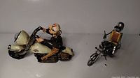 Two metal motorcycle figurines, one rider on white motorcycle and one skeleton rider on different bike, displayed on a plain surface.