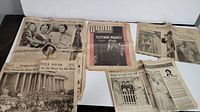 View of seven different old newspapers laid out showing various headlines and photos including mourning events and historical ceremonies.