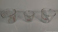 Three glass measuring cups: one 1-cup Fire King, one 2-cup Fire King, and one Pyrex measuring cup, all clear glass with red measurement markings and spouts.