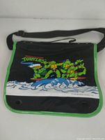 Front view of Teenage Mutant Ninja Turtles vintage duffle bag showing the colorful TMNT 1989 design and logo on black flap with green trim.
