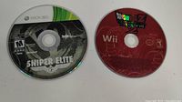 Two game discs: left is Sniper Elite V2 for Xbox 360, right is Dragon Ball Z: Budokai Tenkaichi 2 for Nintendo Wii, laying flat on white surface.