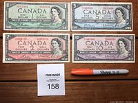 All four banknotes laid out on a wooden table alongside an orange Sharpie for scale: $1 green, $2 pink/black, $5 blue, $10 purple.