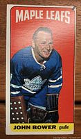 Front of the 1964-65 Topps John Bower Tall Boy hockey card with red background and image of John Bower in Toronto Maple Leafs uniform.