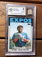 Front view of the 1986 Topps Tim Raines baseball card in protective casing showing the player's image and embedded piece of game used bat with BCCG Near Mint 9 grading label.