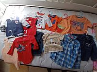 Photo showing various boys' tops and onesies with graphics and designs, including orange t-shirts, denim jacket, plaid shirt, and others laid out on a bed.
