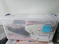 Clear plastic storage crate containing assorted folded curtains in various colors and fabrics.