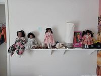 Photo of white shelf holding four dolls dressed in various outfits, a white lamp, and small decorative items including a pink box and toy truck.