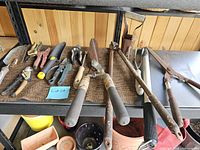 Full view of all gardening tools on shelf showing various handheld implements including pruning shears, trowels, and large loppers.