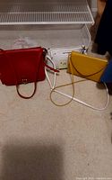 Three purses arranged near a metal shelf: red leather purse, yellow envelope purse, and white zippered pouch.