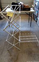 White metal foldable laundry drying rack and white metal shoe rack placed on a garage floor, next to a folding table and storage bins.