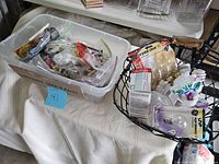 Two baskets on a white cloth. One plastic bin labeled 'Hardware Odds & Ends' full of small packaged hardware pieces, nails, screws, and fittings. A wire basket contains a brass handle, light bulbs in packaging, and other hardware.