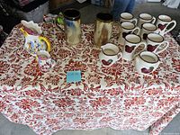 Full lot view on table with red floral tablecloth, showing all mugs, creamers and pitchers.