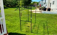 Black metal outdoor frame situated on green lawn next to a porch with white railings. The frame shows a hanging hook where a chime could be attached (chime not included).
