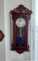 Front view of pendulum wall clock showing carved wood case and clock face with pendulum visible.