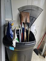 Image showing large black plastic trash can filled with assorted yard tools including shovels and rakes. Broom with red handle leaning against the wall nearby.