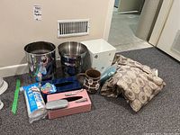 Photo showing various bathroom supplies including metal and MDF wastebaskets, folded mat, Kleenex holders, ceramic pitcher, stapler, cotton rounds, soap dish, and shower curtain liner with rings