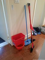 Full view of two red-handled brooms, blue mop, red dustpan, red rectangular bin, and red round basin grouped together on wood floor near wall.