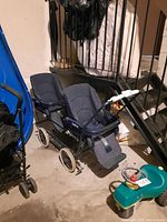 Side view of Peg Perego double stroller showing two adjacent seats with padded blue fabric and a subtle dotted pattern, white wheels, metal frame, and storage basket underneath.