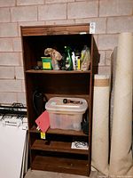 Wooden shelving unit holding cleaning liquids, fire extinguisher, plastic storage container, and rolled material next to it.