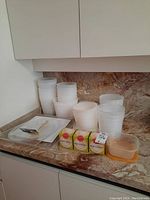 Multiple stacks of semi-translucent white plastic food storage containers with lids, two white square plates, clear plastic forks, and three boxes of parchment baking cups on a kitchen counter.