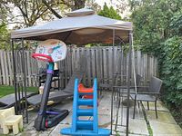 Metal gazebo with brown fabric top on black metal frame, situated on cement pavers with outdoor patio chairs around and children's slide and basketball toy nearby.