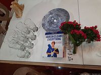 Photo showing 8 clear water glasses, blue swirl glass bowl from Poland, book 'Queen Cleans Everything', faux red rose bouquet and clear glass cake stand plate on a white surface