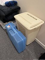 Side view of cream-colored plastic laundry hamper with vented sides and removable lid next to a blue Rubbermaid snap case storage bin on carpeted floor.