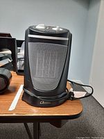 Front view of black portable electric heater with mesh grill and control knobs on top, placed on wooden surface.