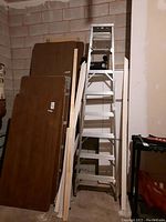 Full view of 7 ft aluminum step ladder standing upright in garage with wood panels leaning against the wall to the side and a shelving unit on the right.
